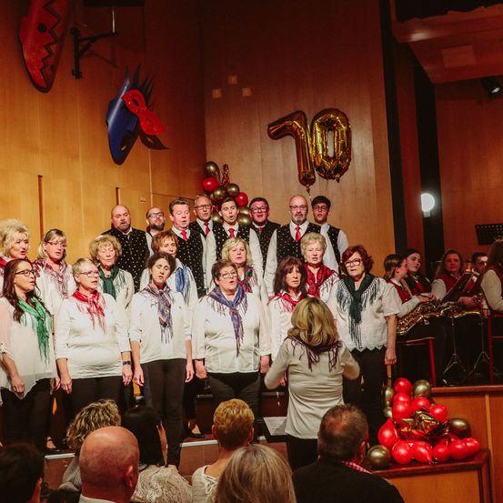 Ein Chor aus Frauen und Männern tritt auf der Bühne auf, mit einer großen Zahl 10 Ballon dahinter. Sie singen, während ein Publikum von vorne zuschaut.