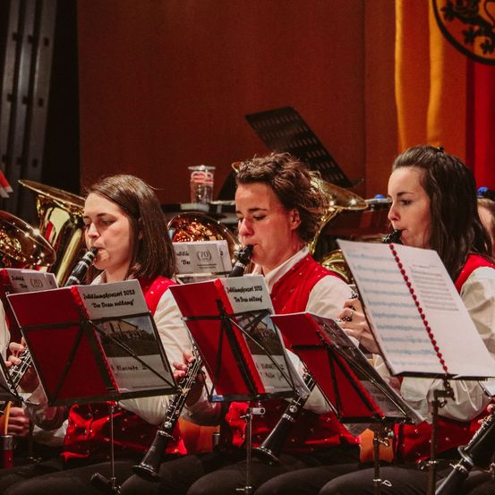 Drei Frauen spielen Klarinette in einem roten Outfit. Sie schauen auf ihre Notenblätter vor sich. Dahinter steht ein Blechinstrument.