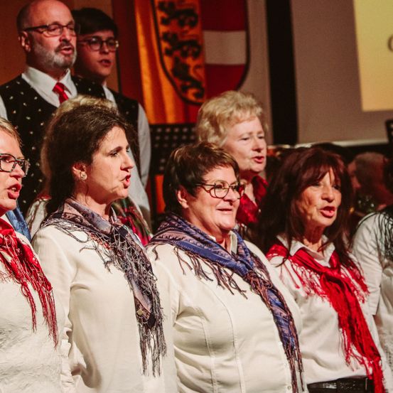 Ein Chor von Frauen singt auf der Bühne, alle tragen weiße Hemden und Schals, mit einer Fahne und anderen Menschen im Hintergrund.