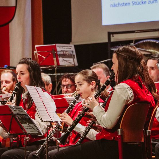 Eine Gruppe von Musikern in roten Westen spielt Klarinetten bei einem Konzert, mit Notenblättern vor ihnen und einer Projektionsleinwand im Hintergrund.