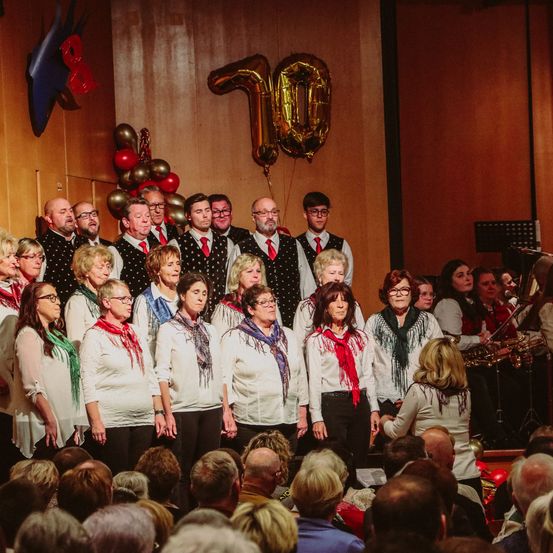 Ein Chor von Frauen und Männern in Weiß mit roten Schals singt auf der Bühne, mit Musikinstrumenten dahinter, einschließlich eines Luftballons in Form der Zahl 10.