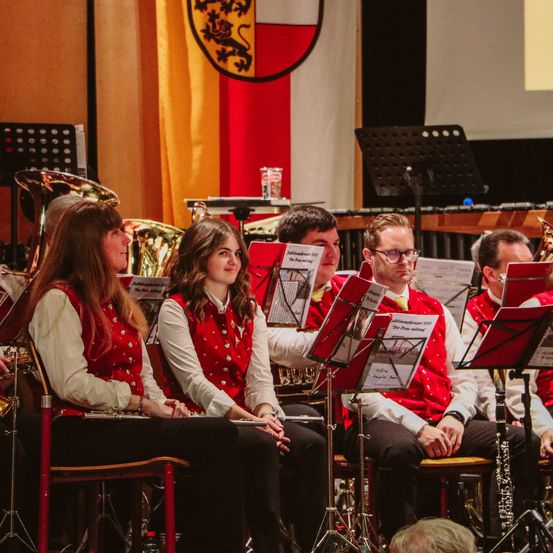 Eine Gruppe von Musikern in roten Westen sitzt auf Stühlen und spielt Instrumente, einige halten Notenständer. Der Hintergrund zeigt eine Wand mit einer rot-weißen Flagge und einem Banner.
