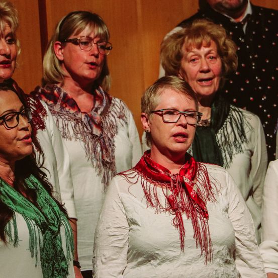 Fünf Frauen, alle mit Brille, stehen zusammen, wahrscheinlich in einem Chor, wobei eine Frau ein rotes Tuch und eine andere ein grünes Tuch trägt. Sie scheinen zu singen oder aufzutreten. Dahinter ist ein verschwommener Hintergrund von einem Mann, der eine gepunktete Weste trägt.