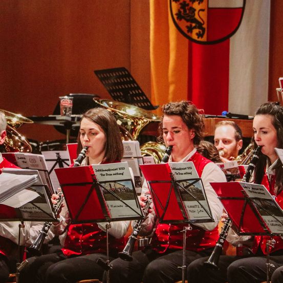 Eine Gruppe von Musikern spielt Klarinetten, während sie in einer Reihe sitzen. Sie tragen rote Westen. Ein Flagge ist im Hintergrund zu sehen.