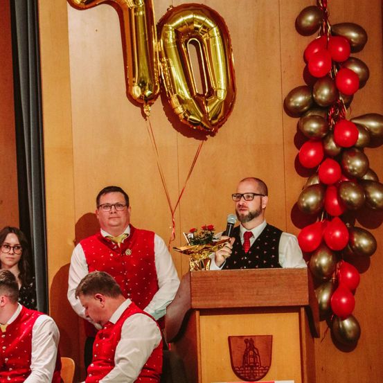 Ein Mann am Rednerpult spricht in ein Mikrofon, während zwei Männer in roten Westen hinter ihm sitzen. Hinter dem sprechenden Mann befindet sich ein goldener Ballon mit der Aufschrift '10'.