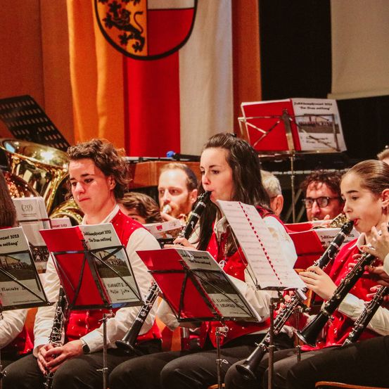Vier Musiker spielen Klarinette und Posaune und lesen Noten, tragen rote Westen und weiße Langarmhemden. Sie sitzen vor einem Musikständer mit roter Abdeckung.