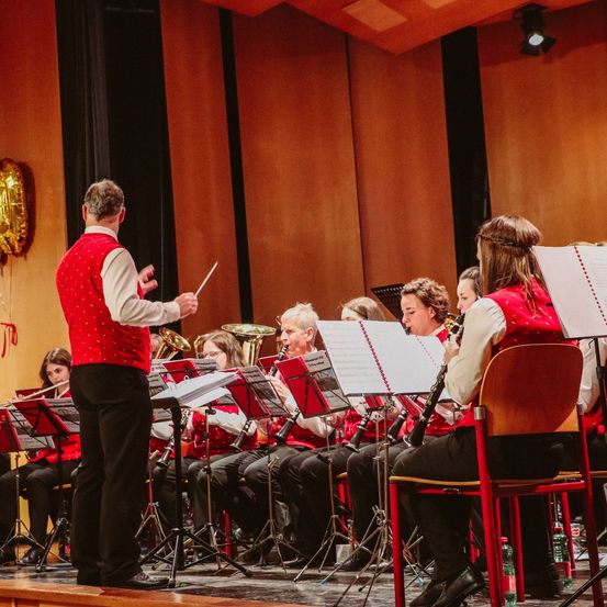 Ein Orchester mit einem Dirigenten tritt auf der Bühne auf. Der Dirigent hält einen Taktstock und trägt eine rote Weste. Die Orchestermitglieder sitzen und stehen und spielen ihre Instrumente.