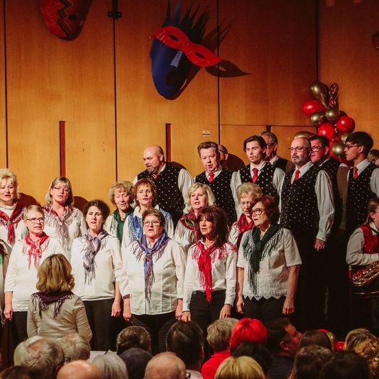 Ein Chor von Frauen und Männern, gekleidet in weißen Hemden, steht auf der Bühne vor einem Publikum. Dahinter befindet sich eine Holzwand, geschmückt mit einer roten Maske und Luftballons.