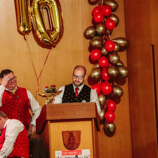 Drei Männer stehen an einem Rednerpult, einer spricht, zwei andere lauschen. Dahinter ein goldener Ballon mit der Nummer 10 und eine Säule aus roten und goldenen Luftballons.