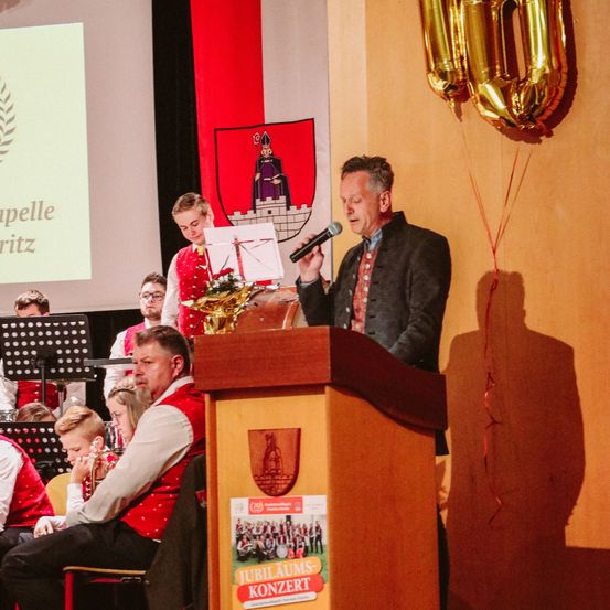 Ein Mann hält eine Rede an einem Podium mit Mikrofon in einem Raum mit einem Orchester. Hinter ihm steht ein großes Plakat und Luftballons. Einige Leute sitzen auf Stühlen vor dem Podium. Ein Junge hält eine Trompete. Ein junges Mädchen steht vor der Bühne.