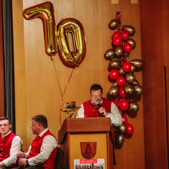 Ein Mann spricht in ein Mikrofon an einem Podium, über dem ein goldener Zahlballon mit der Nummer 10 hängt.