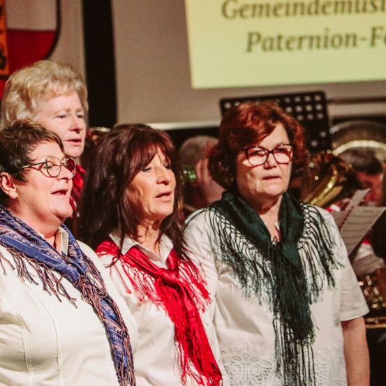 Vier erwachsene Frauen stehen bei einer Musikveranstaltung nebeneinander. Die Frau links trägt ein blaues Tuch. Die Frau in der Mitte trägt ein rotes Tuch. Die Frau rechts trägt ein grünes Tuch. Hinter ihnen sind Musikinstrumente und ein Banner mit den Worten 'Gemeindemusik' und 'Paterniion-Fe' zu sehen.