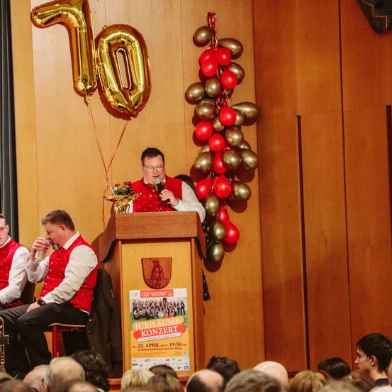 Ein Mann hält eine Rede an einem Podium mit einem goldenen Luftballon, der '10' steht. Er hält ein Mikrofon in der Hand. Zwei andere Männer sitzen hinter ihm auf einem Stuhl, einer von ihnen trinkt Wasser. Ein großes Publikum beobachtet sie.