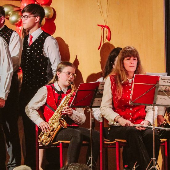 Eine Schulband spielt Instrumente auf der Bühne. Die Saxophonistin sitzt auf einem Stuhl, und die Flötistin ist neben ihr. Hinter ihnen steht ein Trompeter. Die Wand hinter ihnen ist mit Luftballons dekoriert.