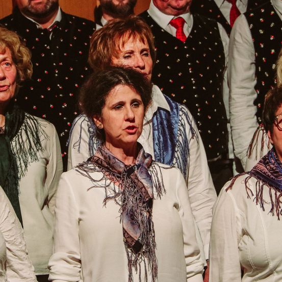 Eine Gruppe von Menschen in weißen Hemden und Westen, wobei die Frauen Schals tragen, steht zusammen und scheint zu singen. Der Hintergrund ist unscharf, aber es sieht so aus, als wären sie in einem Raum mit Holzwänden.