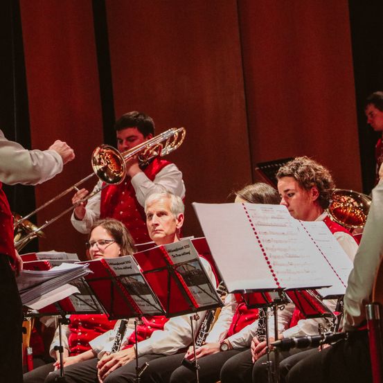 Eine Band spielt Instrumente auf der Bühne bei einem Konzert. Ein Mann spielt die Posaune, und mehrere andere sitzen mit Musikpartituren vor sich.