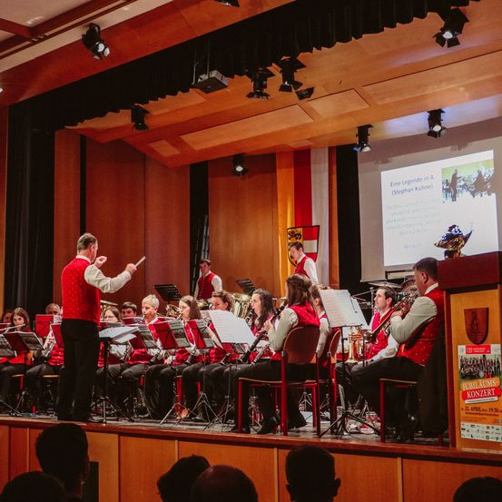 Eine Blechbläsergruppe auf der Bühne gibt ein Konzert für ein Publikum in einem Raum mit einer Holzwand und einer Projektionsleinwand.