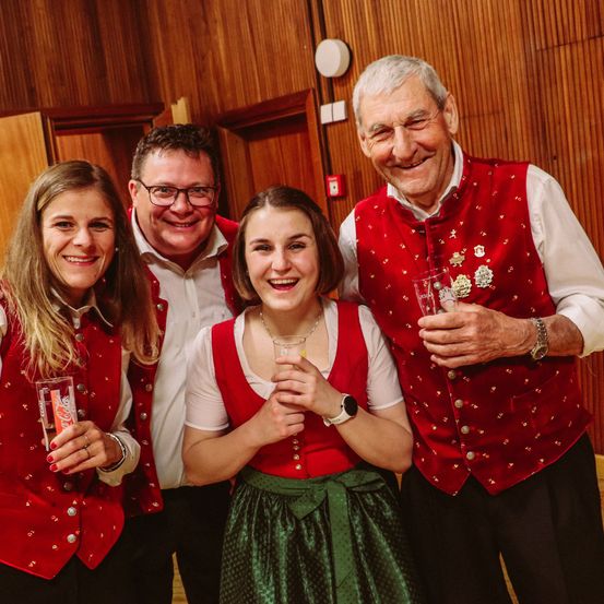 Vier Menschen lächeln und posieren für ein Foto in einem Raum. Zwei Frauen und zwei Männer, alle in traditioneller bayerischer Kleidung. Die beiden Frauen halten Coca-Cola-Gläser.