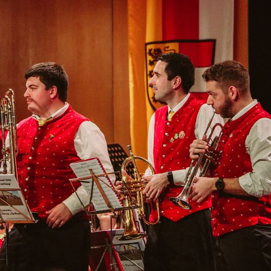 Vier Musiker spielen bei einem Konzert. Drei von ihnen halten Trompeten und sind in roten und weißen Trachten gekleidet. Die vierte Person schaut auf ein Musikblatt auf einem Ständer. Dahinter ist eine Flagge mit einem Logo.