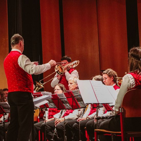 Ein Dirigent in einem roten Weste leitet ein Orchester bei einem Konzert. Die Musiker sitzen vor ihm und spielen verschiedene Instrumente.