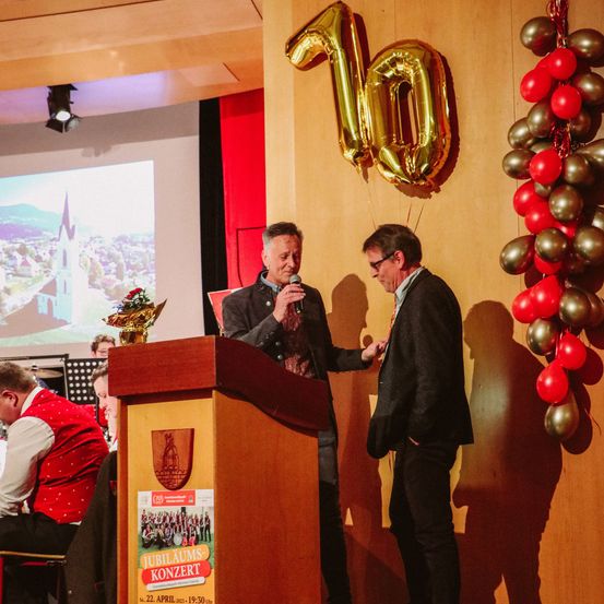 Zwei Männer stehen auf einer Bühne mit einem goldenen Luftballon, der 10 sagt, einem großen Plakat und Luftballons an der Wand.