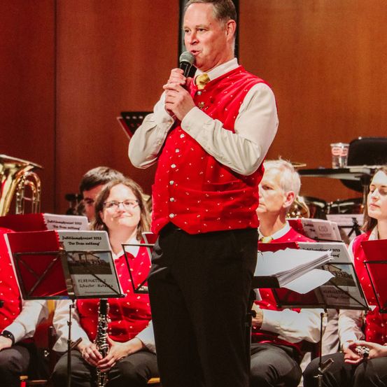 Ein Mann mit Mikrofon steht vor einer Gruppe von Musikern in roten Westen. Die Musiker spielen verschiedene Instrumente, darunter Tuba, Klarinette und Flöte.