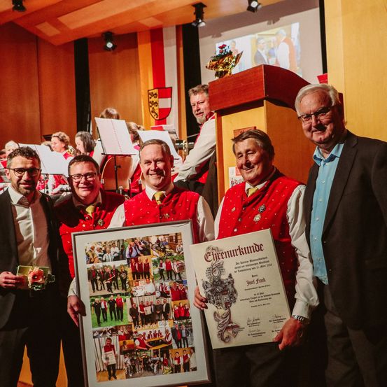 Fünf Männer stehen in einer Reihe mit lächelnden Gesichtern. Sie halten Urkunden und eine gerahmte Fotocollage in der Hand. Einer der Männer hält eine goldene Trophäe. Sie befinden sich in einem Saal mit Bühne und einer großen Leinwand hinter ihnen.