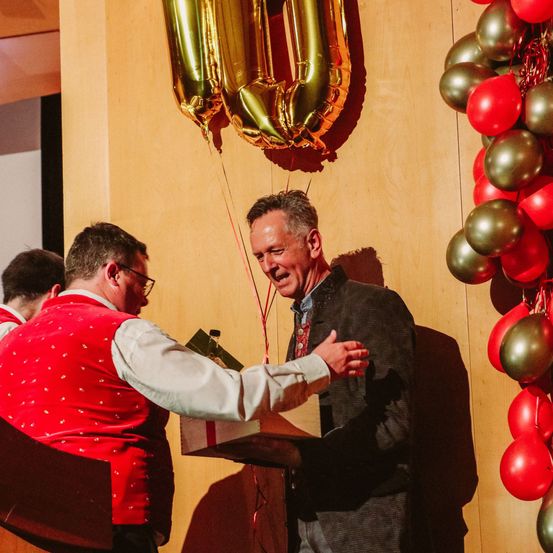 Ein Mann überreicht einem anderen Mann ein Geschenk, während er neben einem Podium steht. Sie stehen neben einer Wand mit Luftballons und der goldenen Zahl 100.