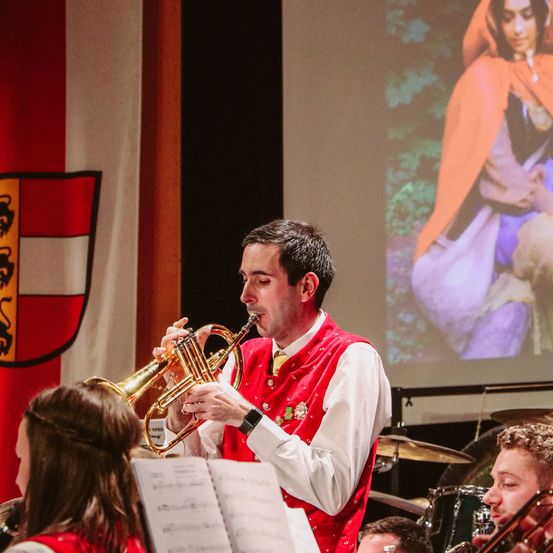 Ein Mann spielt Trompete in einer Band, während eine Frau zusieht. Eine Frau in einem roten Umhang wird auf der Leinwand hinter ihm projiziert.