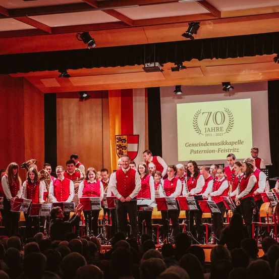 Ein Musikband tritt auf der Bühne auf, vor ihnen ein Publikum, ein Mann steht vor der Gruppe und hinter ihnen ein großer Bildschirm mit Text.