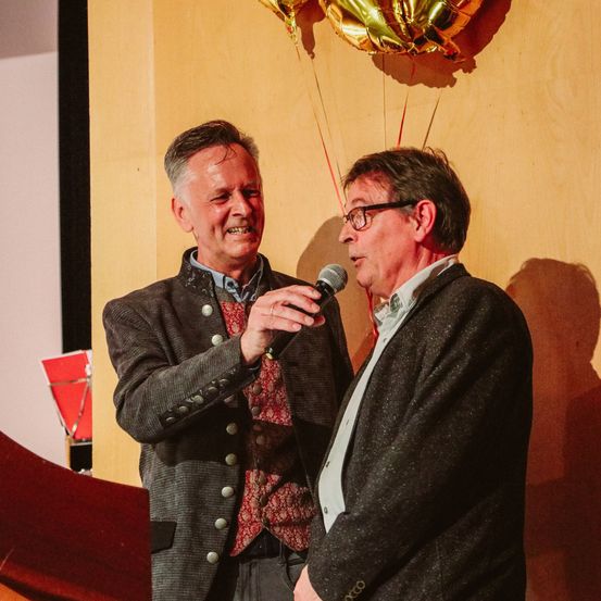 Zwei Männer stehen neben einem Podium mit einem Mikrofon, einer hält es und scheint zu sprechen, während der andere zuhört. Hinter ihnen ist eine Wand mit einem goldenen Luftballon, der daran hängt.