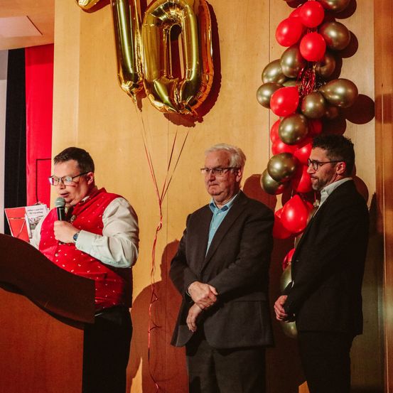 Ein Mann in einer roten Weste steht hinter einem Podium, während zwei Männer in Anzügen neben ihm stehen, mit einem großen goldenen Luftballon mit der Nummer 10 und einer Ansammlung roter und goldener Luftballons im Hintergrund.