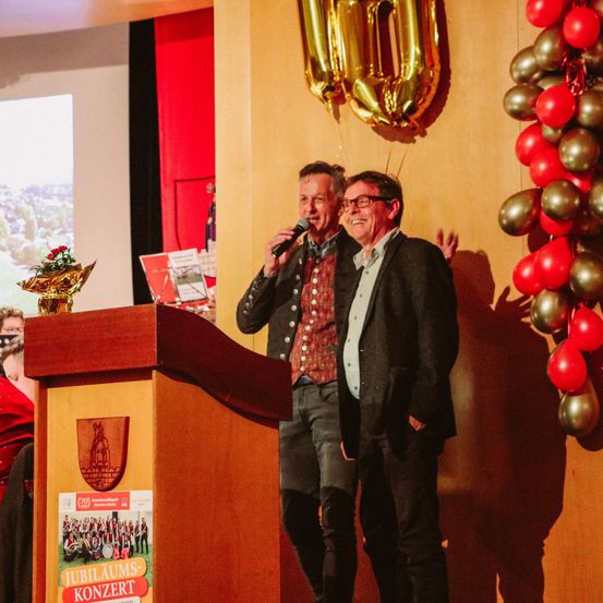 Zwei Männer auf der Bühne während eines Konzerts, einer von ihnen hält ein Mikrofon und der andere lächelt. Hinter ihnen eine Wand mit einer goldenen Nummer 10, roten und goldenen Luftballons und einem Podium mit einem Plakat, das 'Jubiläums-Konzert' liest.