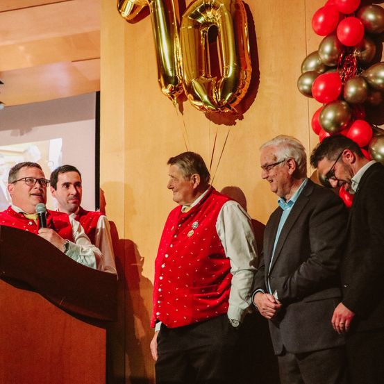 Vier Männer stehen nebeneinander vor einer Wand, zwei von ihnen tragen rote Westen. Einer von ihnen hält ein Mikrofon, wahrscheinlich spricht er. Ein goldener Folienballon mit der Nummer 10 hängt hinter ihnen.