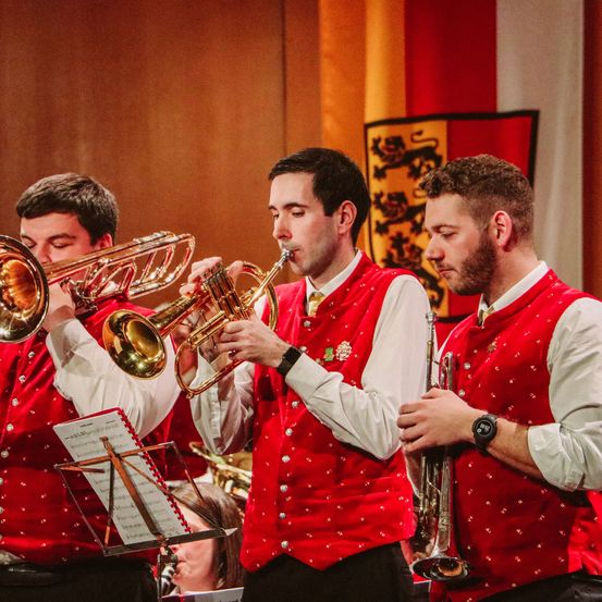 Drei Männer in roten Westen und weißen Langarmhemden spielen Blechblasinstrumente. Der Mann in der Mitte spielt die Trompete. Dahinter ist ein verschwommener Blick auf eine Flagge mit Löwendesign und einem Notenständer mit Notenblättern.