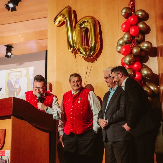 Vier Männer stehen an einem Podium und scheinen zu sprechen. Ein Mann spricht in ein Mikrofon, während die anderen drei zuhören. Hinter ihnen schmücken eine große Zahl 10 und ein Strauß roter und goldener Luftballons die Wand.