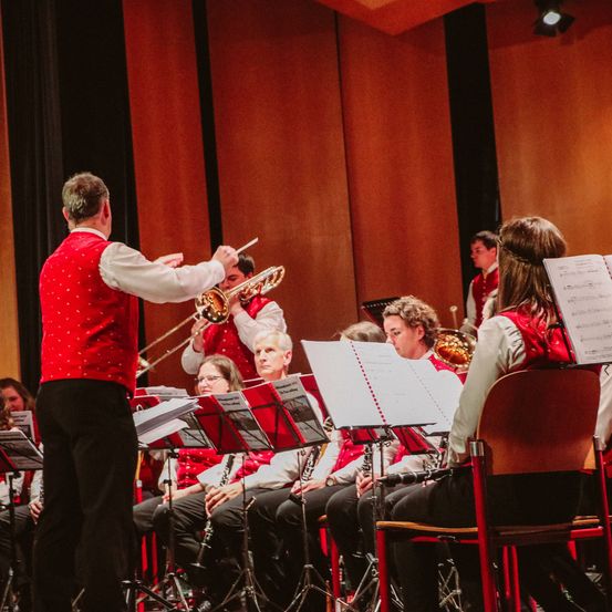 Ein Mann in einem roten Vest spielt Posaune vor einem Orchester, dessen Mitglieder auf roten Stühlen sitzen.