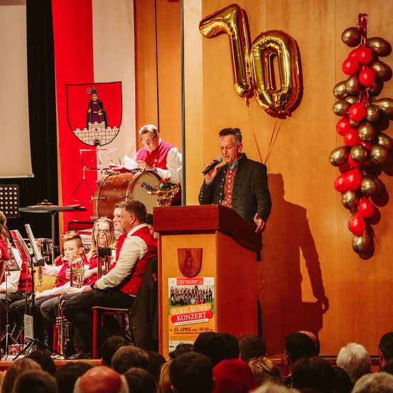 Ein Orchester tritt bei einer Veranstaltung auf. Ein Mann spricht in ein Mikrofon an einem Podium. Ein Schild am Podium zeigt 'Jubiläumskonzert'. Ein goldener Ballon mit der Nummer 10 schwebt in der Luft.