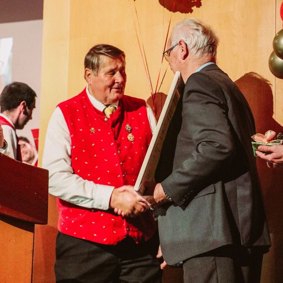 Ein Mann in einem roten Weste erhält eine Auszeichnung von einem älteren Mann im Anzug. Der Mann in der Weste lächelt, während er dem älteren Mann die Hand schüttelt.