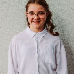 Ein junges Mädchen mit Brille und weißem Hemd lächelt in die Kamera. Es hat eine Tasche auf der linken Seite des Hemdes.