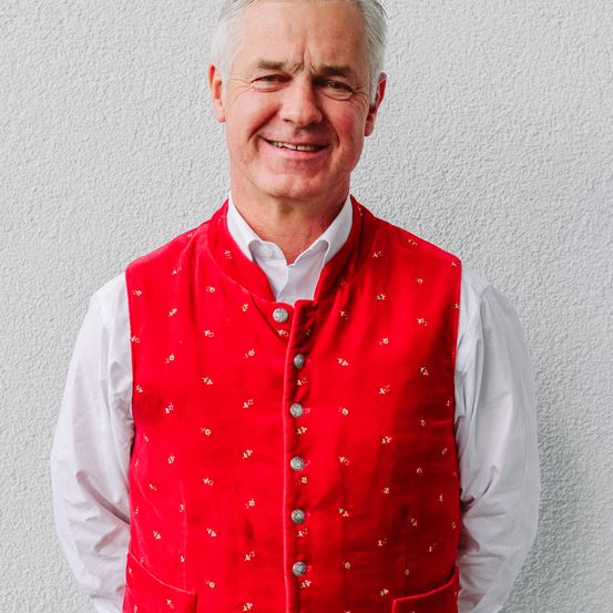 Ein Mann in einem roten Sakko mit Silberknöpfen und einem weißen Hemd steht vor einer weißen Wand.