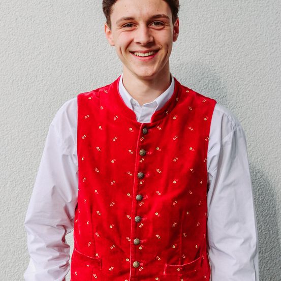 Ein Mann steht vor einer weißen Wand und trägt einen roten Weste mit goldenen Blumenmustern und einem weißen Langarmshirt. Er lächelt.