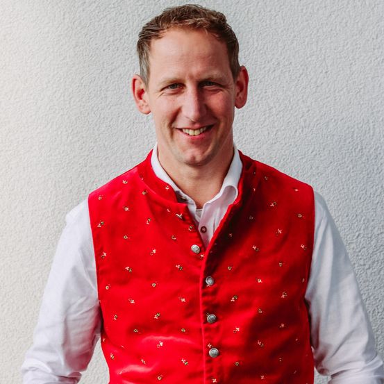 Ein lächelnder Mann trägt ein rotes Blumenweste über einem weißen Hemd vor einer weißen Wand.