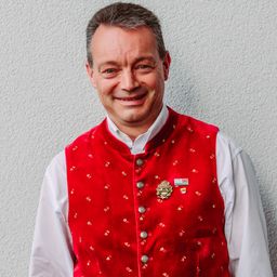 Ein Mann, der eine rote bestickte Weste mit einem weißen Hemd und Knöpfen vorne trägt, lächelt für ein Foto.