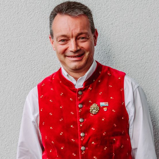 Ein Mann, der eine rote bestickte Weste mit einem weißen Hemd und Knöpfen vorne trägt, lächelt für ein Foto.