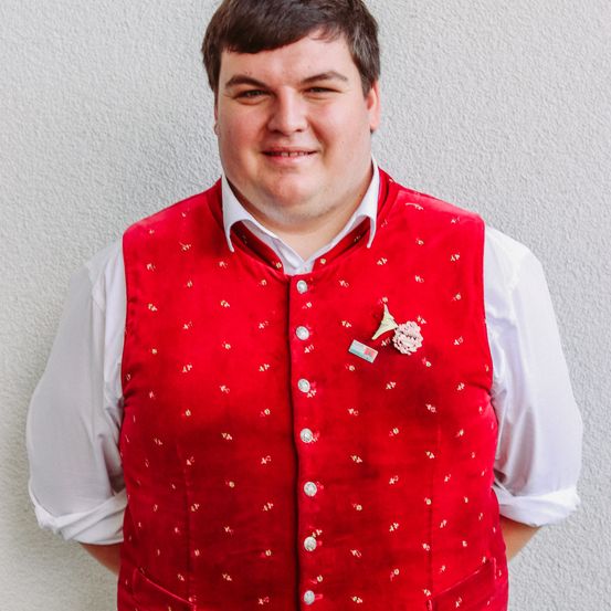 Ein Mann, der eine rote Weste mit goldener Stickerei und ein weißes Innenhemd trägt, steht gegen eine weiße Wand und lächelt in die Kamera.