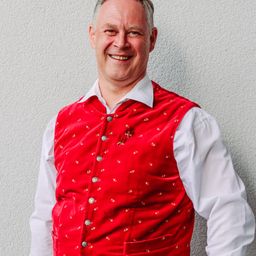 Ein Mann in einem roten Weste mit goldenen Blumen und Knöpfen steht vor einer weißen Wand, lächelt und schaut nach rechts.