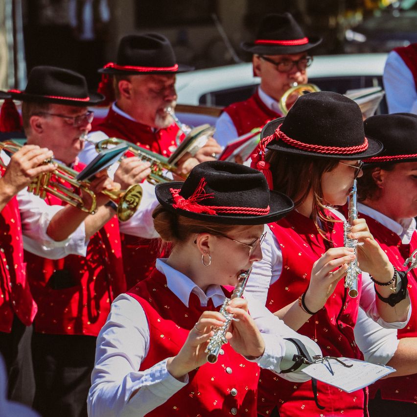 Eine Blaskapelle tritt im Freien auf, wobei Musiker in roten Westen und schwarzen Hüten Trompeten, Flöten und Posaunen spielen. Die Gruppe steht in einer Reihe, wobei einige Mitglieder Musikblätter halten.