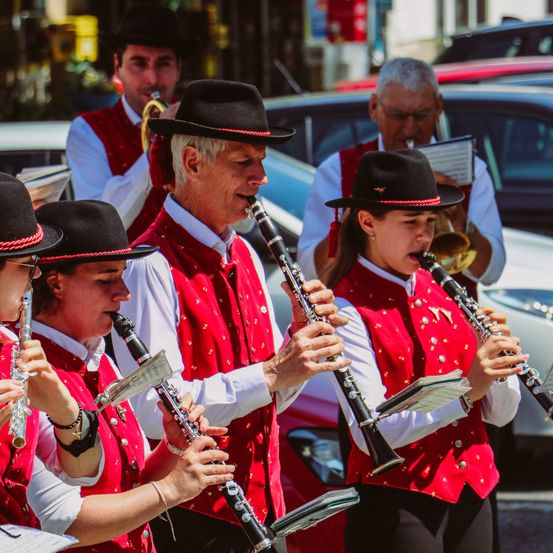 Eine Straßenband von Musikern in roten Westen und Hüten spielt Instrumente. Ein Mann spielt Klarinette, eine Frau neben ihm Trompete. Hinter ihnen spielen andere Bandmitglieder ihre Instrumente. Im Hintergrund sind geparkte Autos.