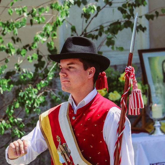 Ein Mann in einem roten Wams und Hut mit einem befransten Stab steht vor einem grünen Baum.